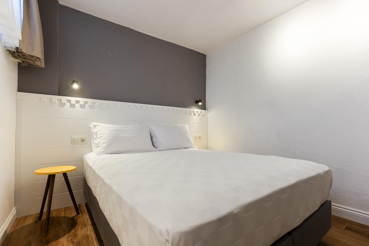 A neatly made bed with white bedding, a small wooden stool, and a wall-mounted light fixture.