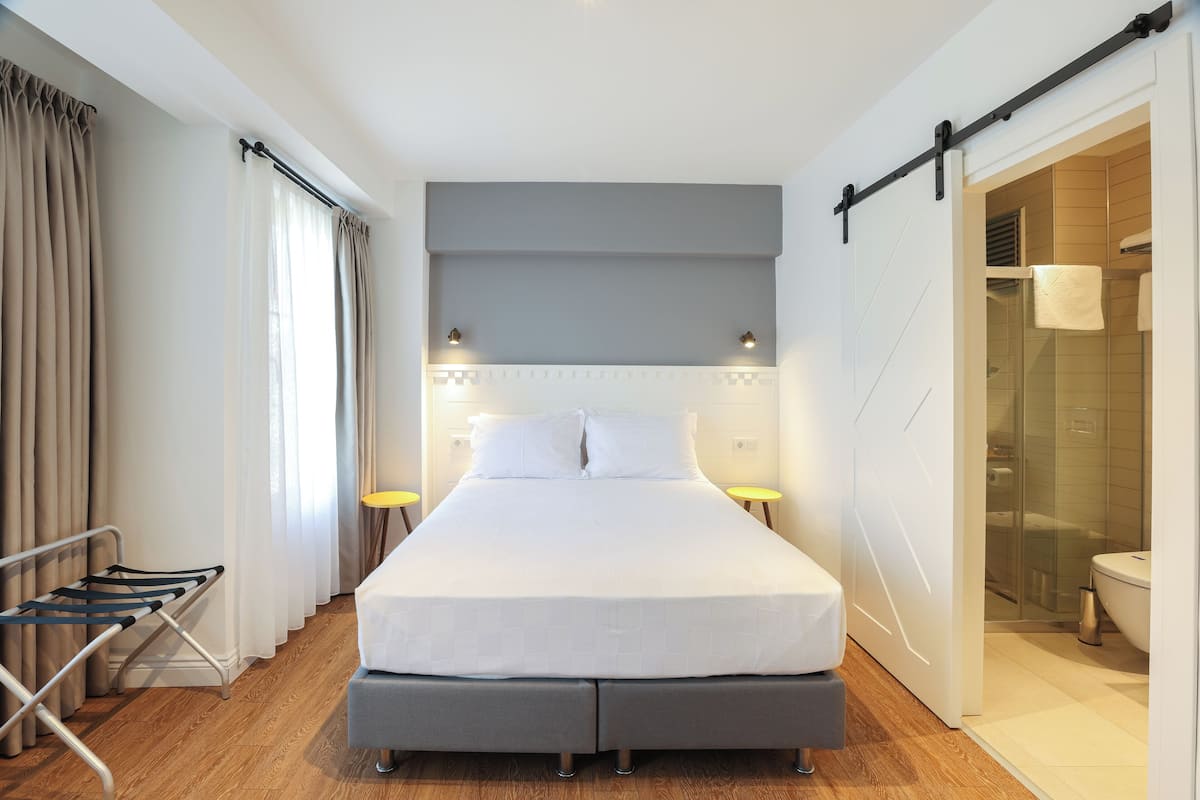 A modern hotel room with a large bed, bedside tables, a built-in shower, and a sliding door to the bathroom.