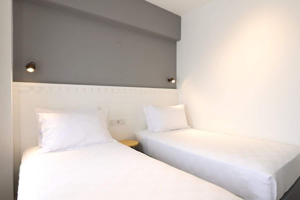 A hotel room with two single beds, a small bedside table, and a light fixture above the beds.