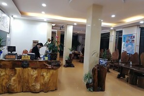 Zona de recepción de un hotel con un mostrador de madera, una computadora y una planta en maceta. Se observan varias sillas y mesas al fondo.