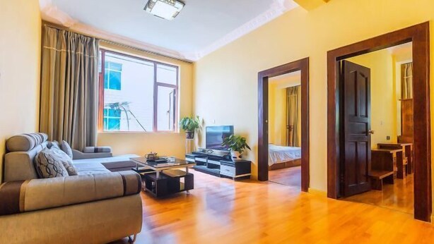 Amplia sala de estar con sofá, una mesa de centro y televisor. Se aprecia un ventanal con cortinas, una puerta que conduce a otra habitación y una planta sobre la mesa de centro.