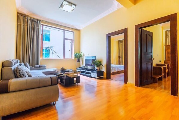 Amplia sala de estar con sofá, una mesa de centro y televisor. Se aprecia un ventanal con cortinas, una puerta que conduce a otra habitación y una planta sobre la mesa de centro.