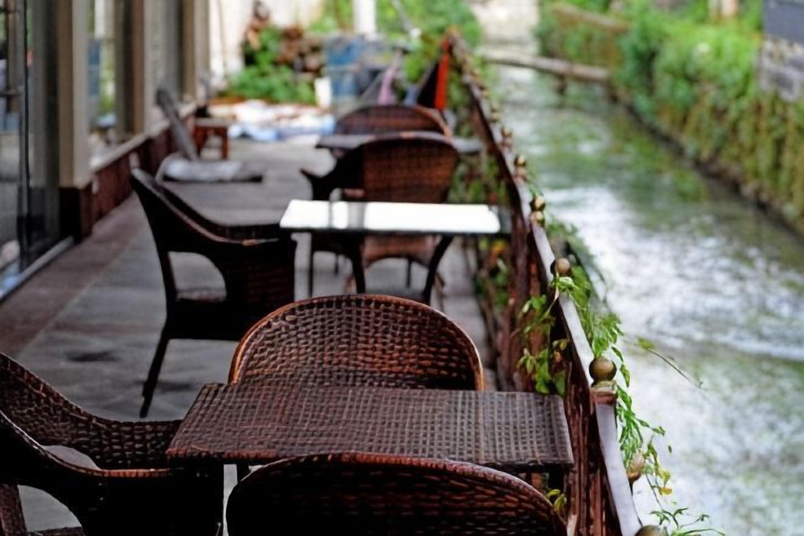 Une rangée de tables et de chaises en osier est disposée dans un espace extérieur abrité, offrant une vue sur un cours d’eau.