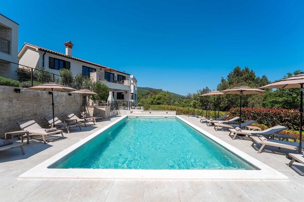 A rectangular swimming pool with lounge chairs and umbrellas, surrounded by a stone wall and a building with multiple balconies.
