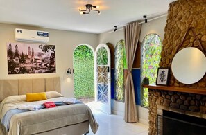 Un dormitorio con cama, pared de piedra, chimenea y un ventanal con vistas a la vegetación.