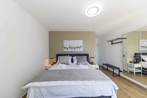 Une chambre à coucher moderne avec un grand lit, une table de chevet, un banc, un miroir et un tableau accroché au mur.