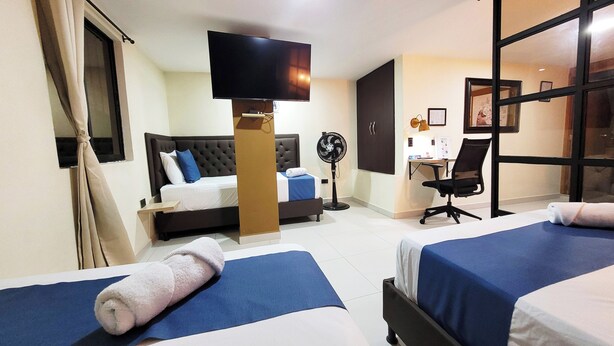 Habitación de hotel con dos camas, televisión, escritorio con silla y ventilador de pie.