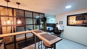 Habitación de hotel compacta con zona de comedor, cama, pequeña kitchenette y un amplio ventanal.