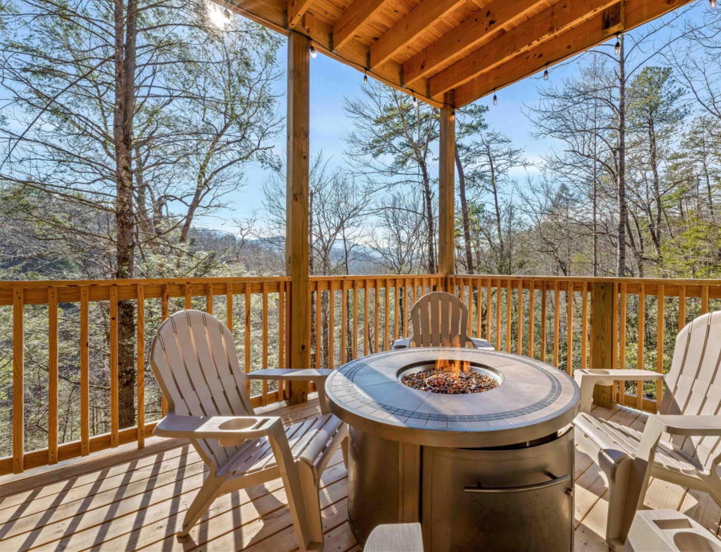 A wooden deck with a round table and chairs, a fire pit, and a view of trees and mountains.