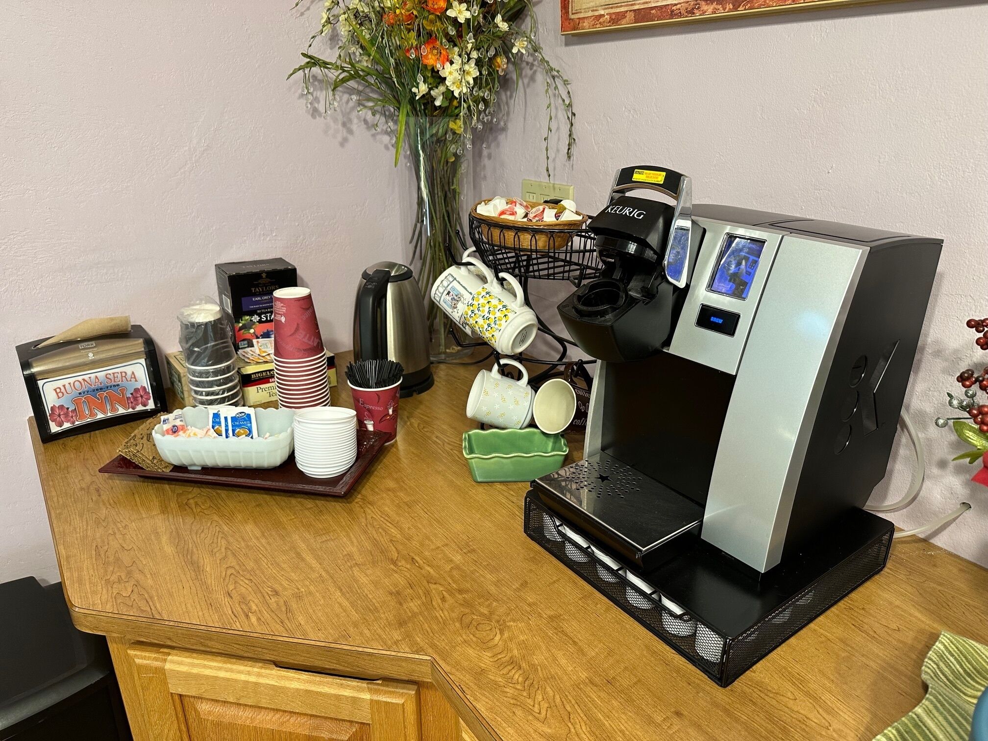 A coffee station with a Keurig machine, disposable cups, a basket of pastries, and a sign reading "Buona Sera Inn."