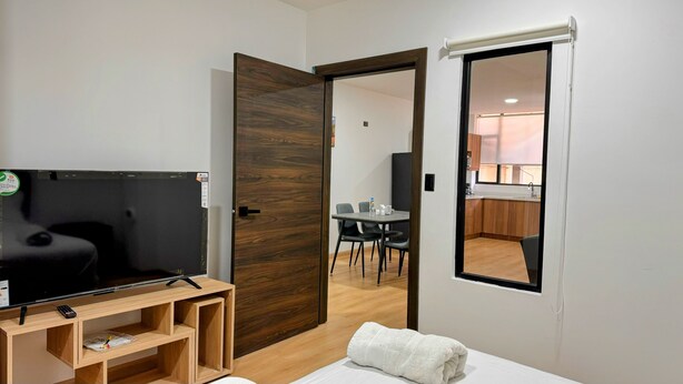 A hotel room with a TV, wooden furniture, a small table, and a bathroom visible through a door. - Urban Serenity: Cozy 1Bedroom apartment. (Quito)