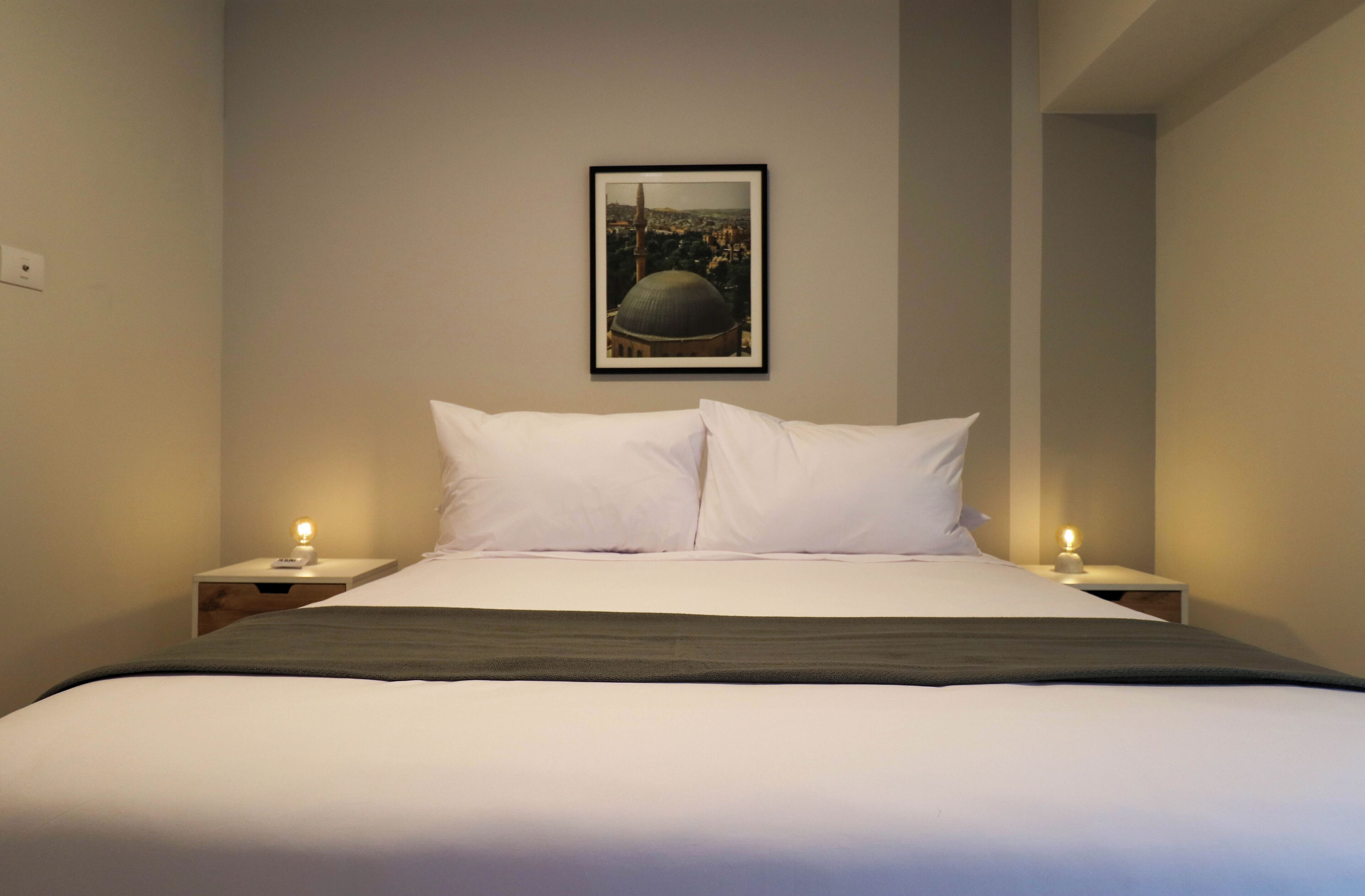 A neatly made bed with white linens and a dark throw blanket, flanked by two bedside tables with lamps. A framed picture of a dome and minaret is hung on the wall above the bed.