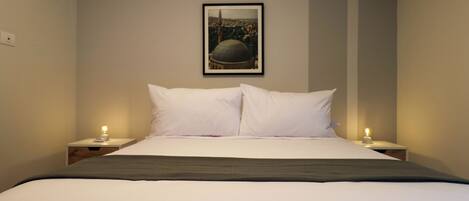 A neatly made bed with white linens and a dark throw blanket, flanked by two bedside tables with lamps. A framed picture of a dome and minaret is hung on the wall above the bed.