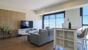 A modern living room with a grey sofa, a flat-screen TV, a dining area with a white table and chairs, and large windows offering a view. - La Mar Miraflores by Wynwood House (Lima)