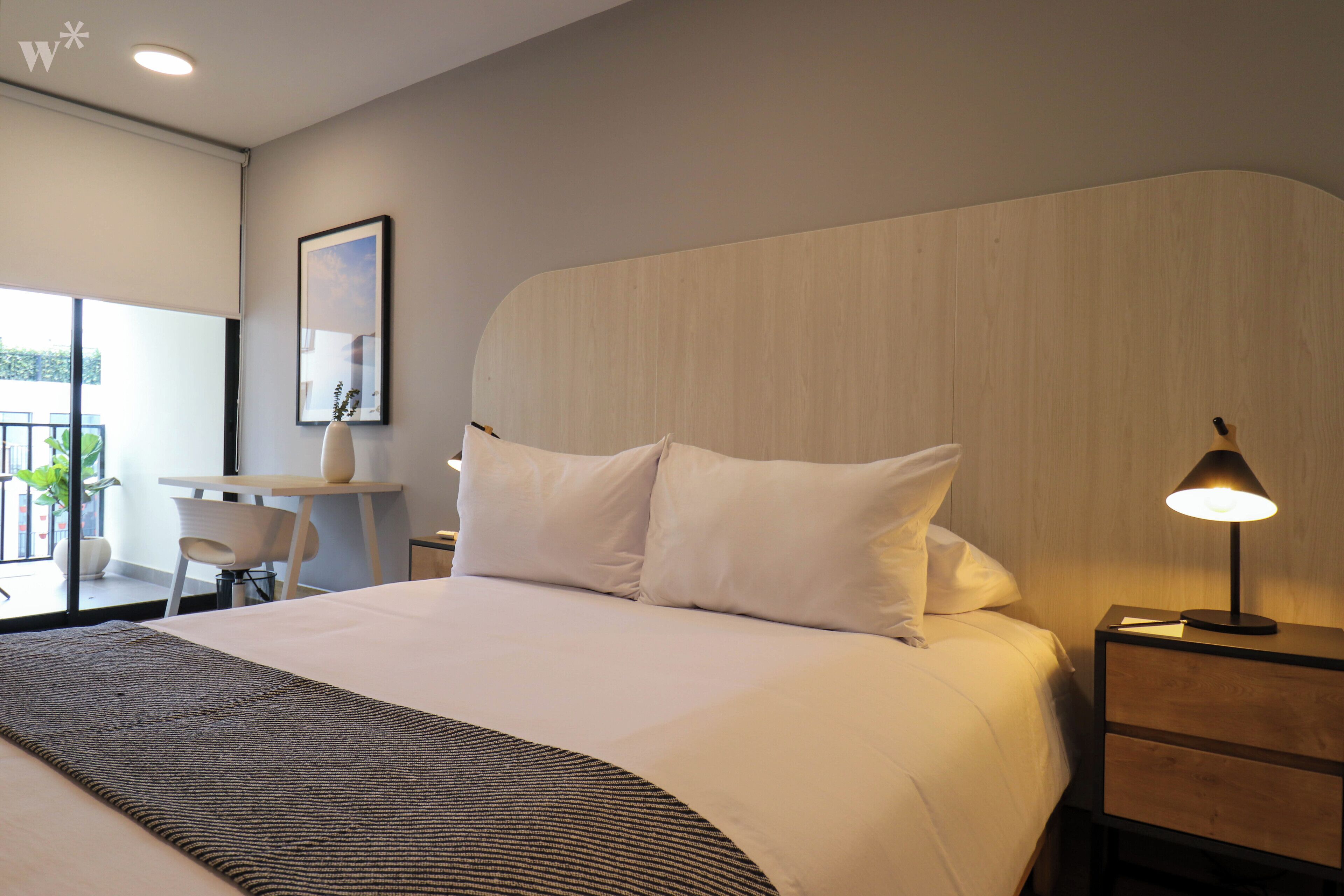 A modern hotel room with a large bed, a bedside table with a lamp, a small desk with a chair, and a window with a view of greenery.
