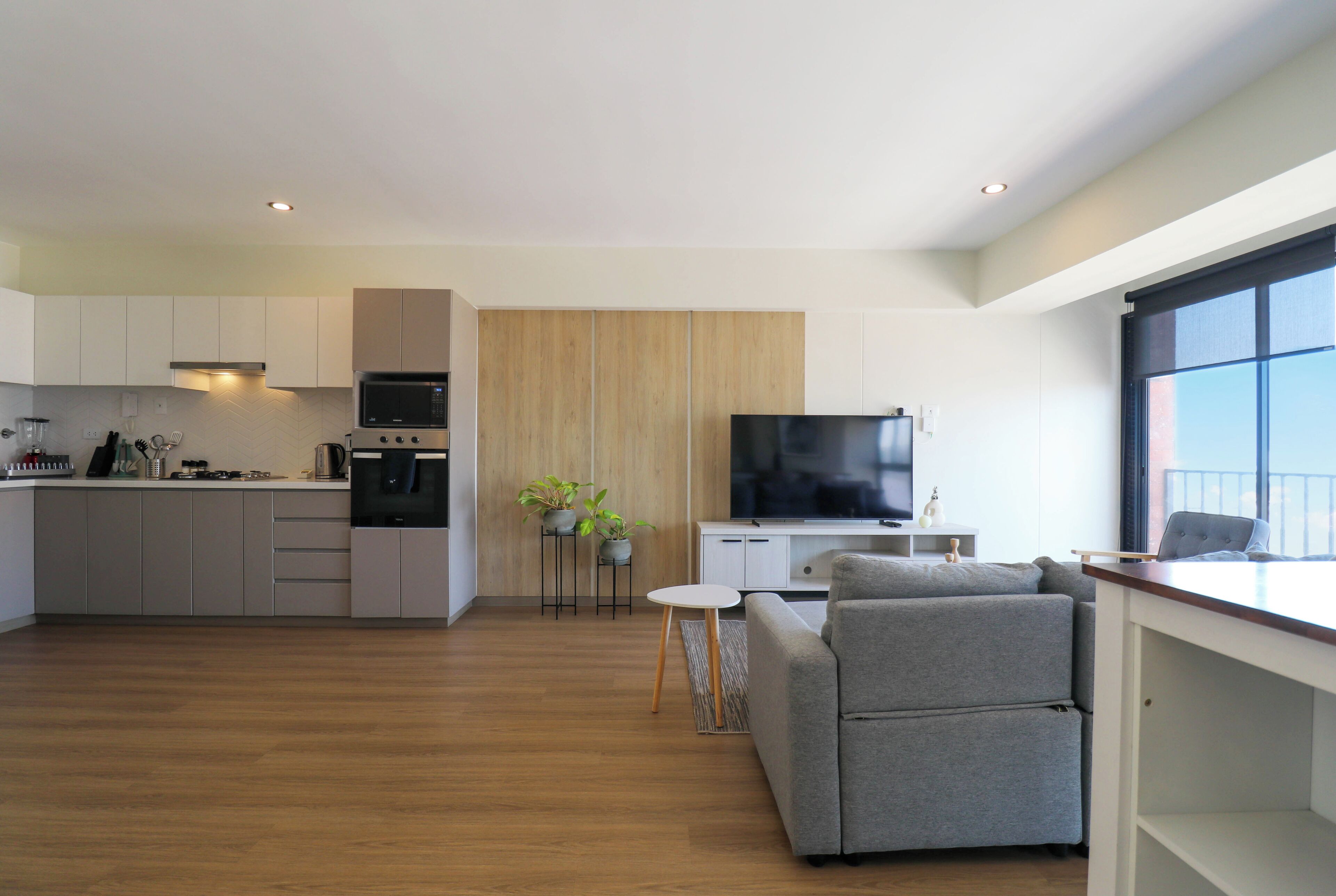 A modern kitchen with a microwave, oven, and sink, a grey sofa, and a TV in a living area with a wooden floor and a large window.