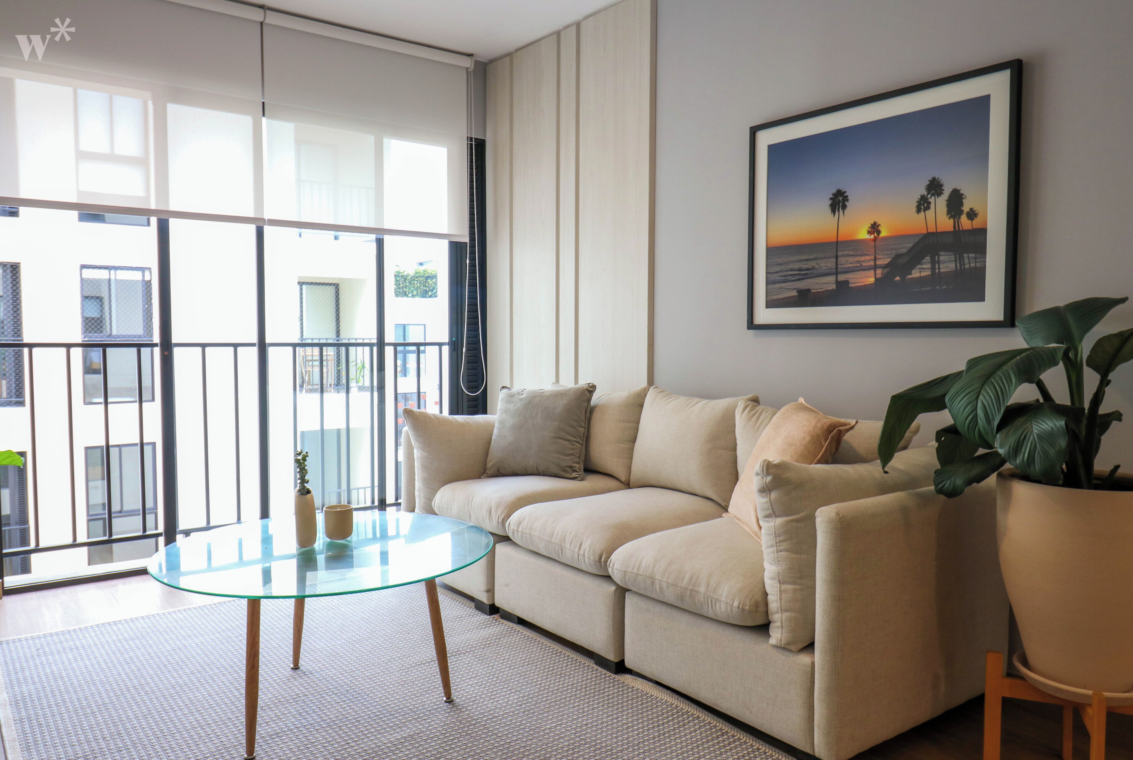 A living room with a glass coffee table, beige sofa, and a framed picture of a sunset beach scene.