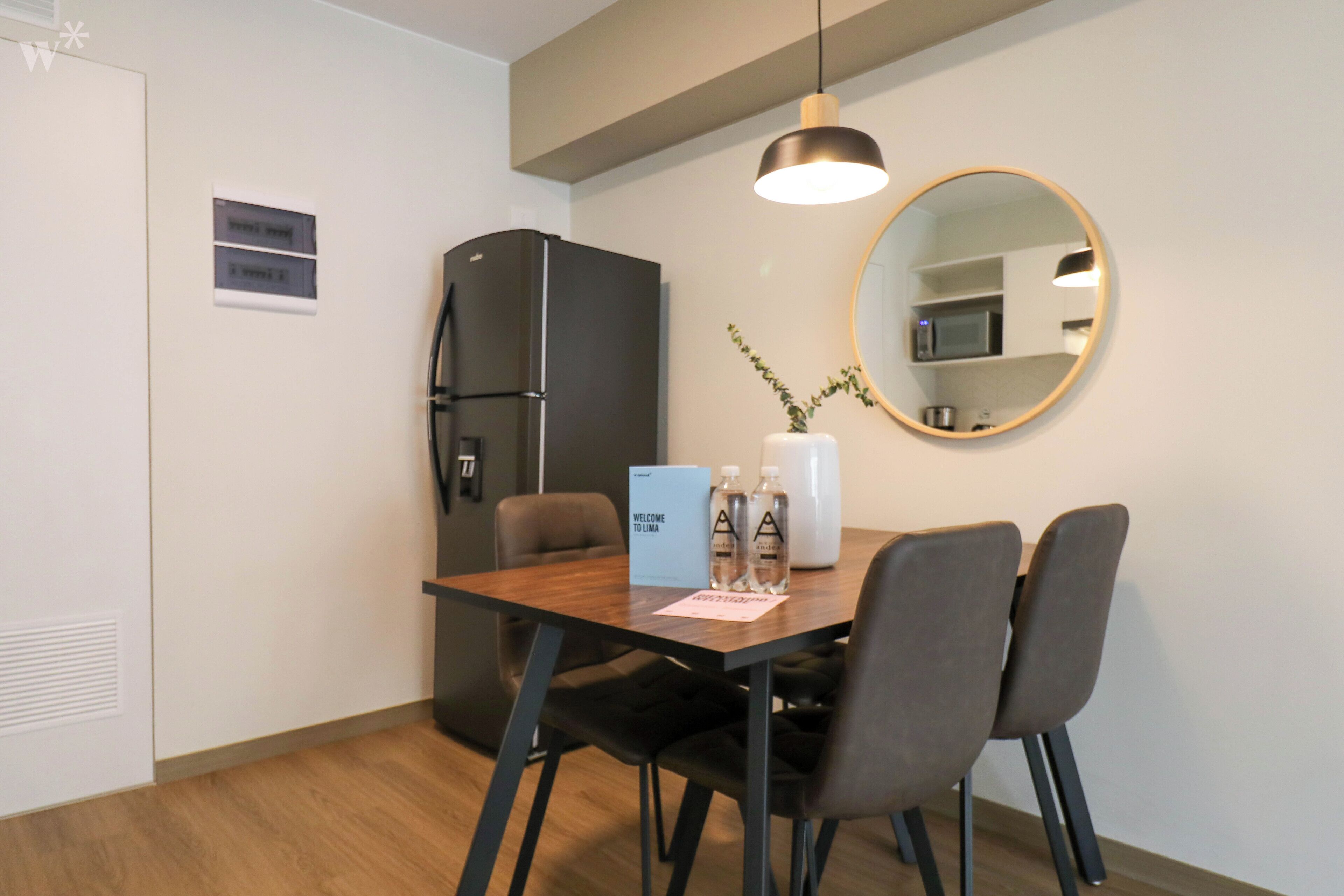 A modern kitchen with a refrigerator, microwave, and a dining table with chairs. There is a welcome sign and a vase with plants on the table.