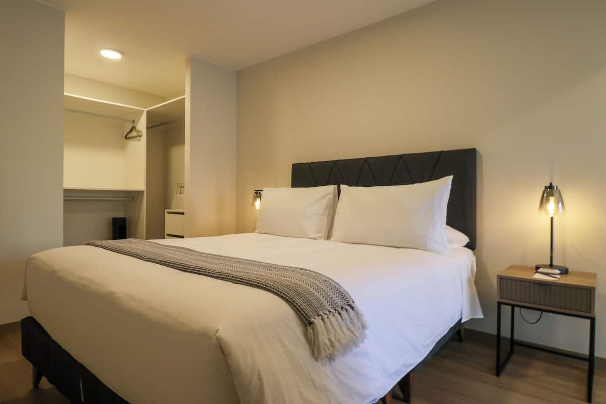 Uma cama bem arrumada com cabeceira, mesa de cabeceira com abajur e um cabideiro.