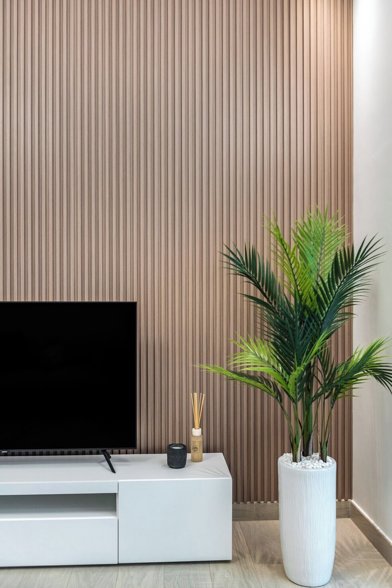 A modern living room with a flat-screen TV, a white entertainment unit, a large potted plant, and a wooden slatted wall.