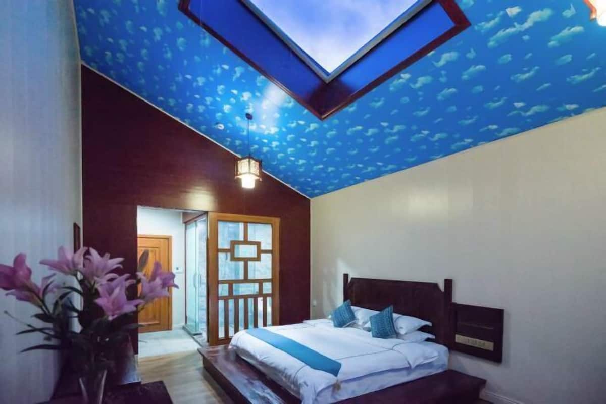A bedroom with a skylight, a bed with pillows, a nightstand, and a vase with flowers.