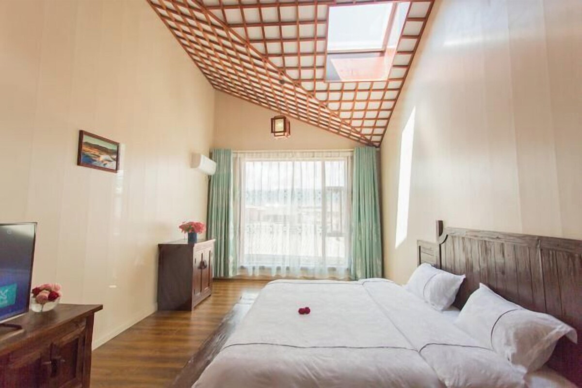A hotel room with a wooden ceiling, a bed with white bedding, a TV on a wooden cabinet, and a window with curtains.