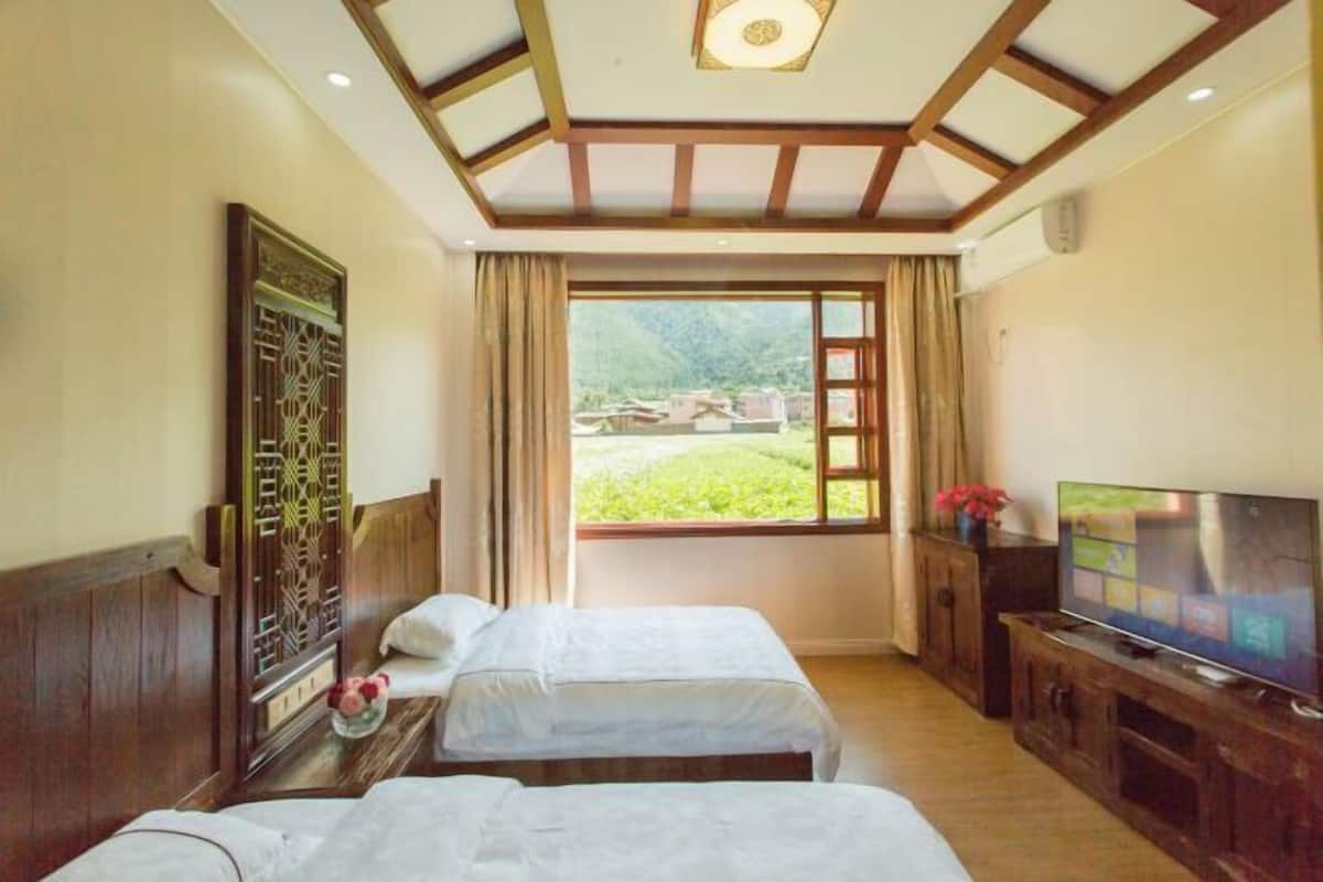 A hotel room with two beds, a TV, a window with a view of greenery, and a decorative wooden panel on the wall.
