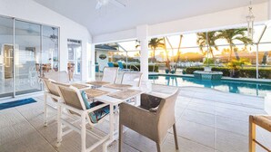 A modern indoor pool area with a glass wall, white furniture, and a view of palm trees and a sunset.