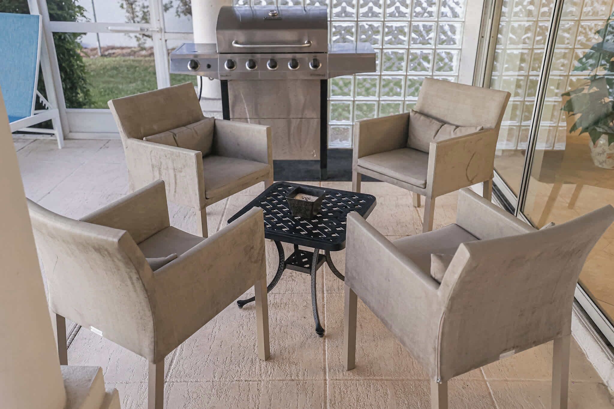 Un área de patio con cuatro sillas color beige y una pequeña mesa, una parrilla de acero inoxidable y una puerta de cristal que da a un jardín.