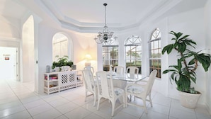 A spacious dining area with a round table, white chairs, and a chandelier. There is a potted plant and a cabinet with decorative items.