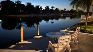 A serene outdoor seating area with a table and chairs by a calm water body, surrounded by palm trees and a clear sky. - Hidden Secret Waterfront Retreat (Cape Coral)