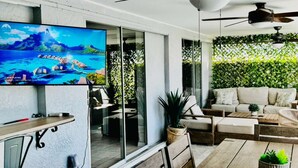 A modern living room with a flat-screen TV, a ceiling fan, and a large window overlooking a lush garden. - Hidden Secret Waterfront Retreat (Cape Coral)