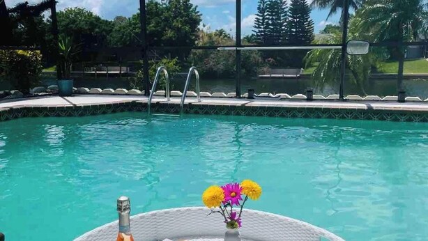 A poolside breakfast setup with a bottle of champagne, a glass of orange juice, a glass of wine, a plate of pastries, a vase with flowers, and a tray with fruits and nuts. - Hidden Secret Waterfront Retreat (Cape Coral)