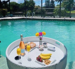 Frühstück am Pool mit einer Champagnerflasche, einem Orangensaftglas, einem Weinglas, einem Teller Gebäck, einer Vase mit Blumen und einer Platte mit Früchten und Nüssen.