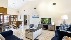 Uma sala de estar ampla com uma televisão grande, assentos confortáveis, uma área de refeições com cadeiras de vime e uma cozinha.