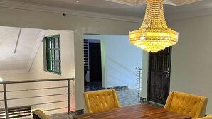 A dining area with a wooden table and four yellow chairs, a chandelier, and a view of a balcony with a railing. - Genisys Guest House (Ijebu Ode)
