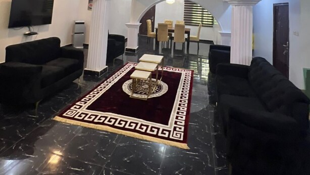 A spacious lobby with a high ceiling, a central rug with a geometric pattern, a television mounted on the wall, and a staircase leading to an upper level. - Genisys Guest House (Ijebu Ode)