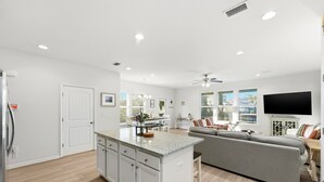 A modern kitchen with white cabinets, a central island, and a dining area with a grey sofa and a flat-screen TV. - Spacious 4BR w/ Pool Access|Walk to Beach|Fire Pit (Port St. Joe)