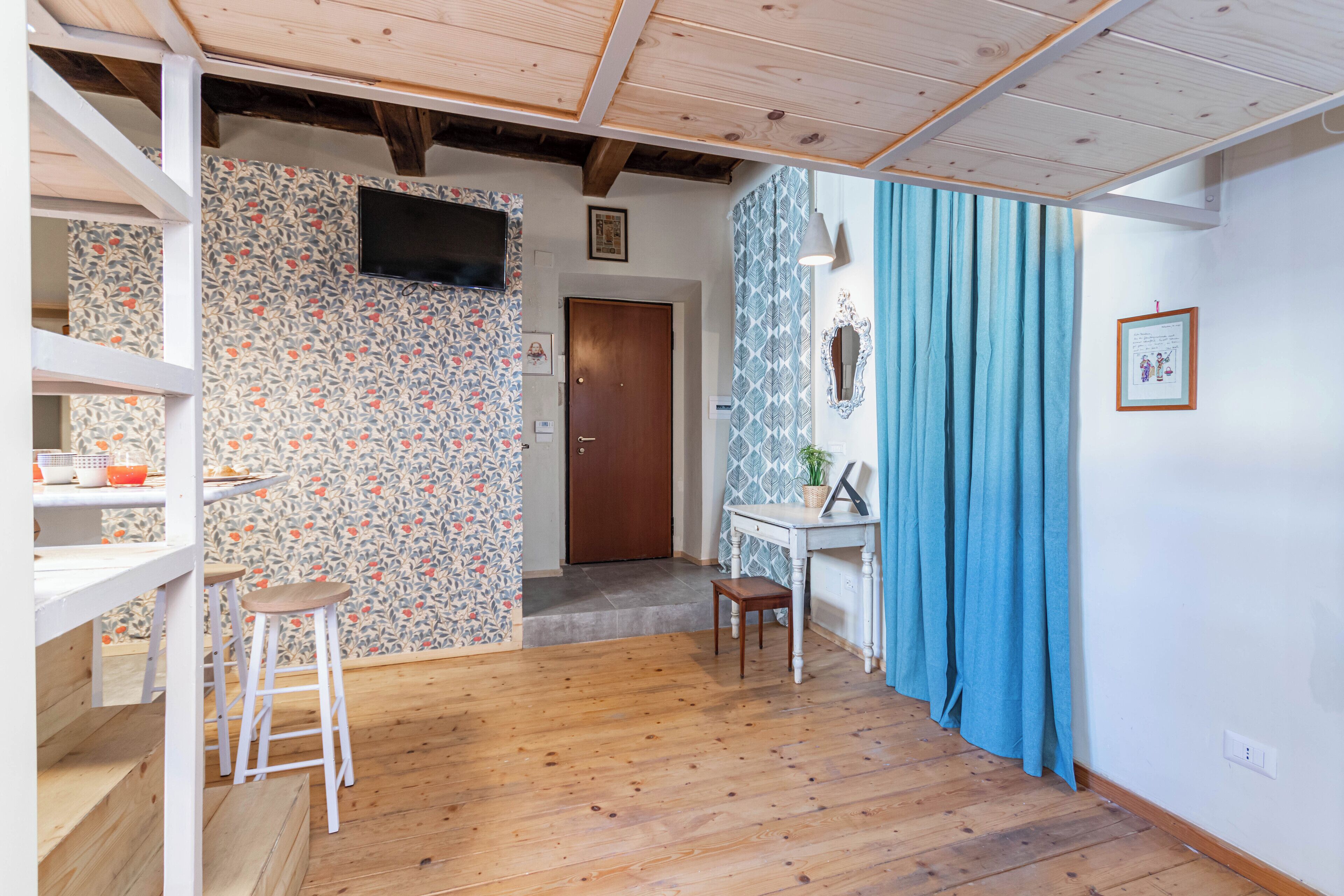 A room with wooden flooring, a white desk with a chair, a television mounted on the wall, and a door leading to another room.