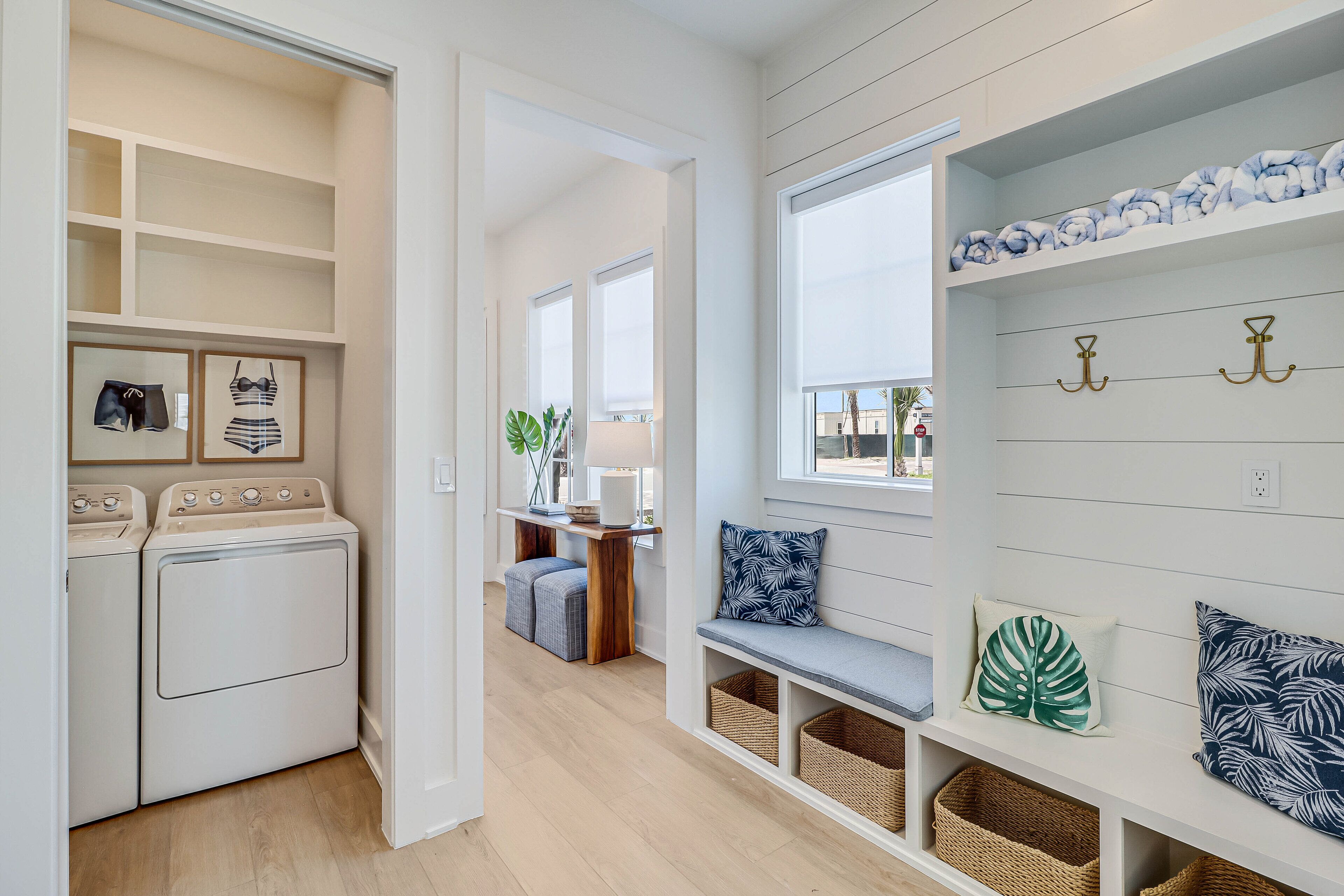 A laundry area with a washing machine and dryer, a bench with storage baskets, and a wall with hooks and decorative pillows.