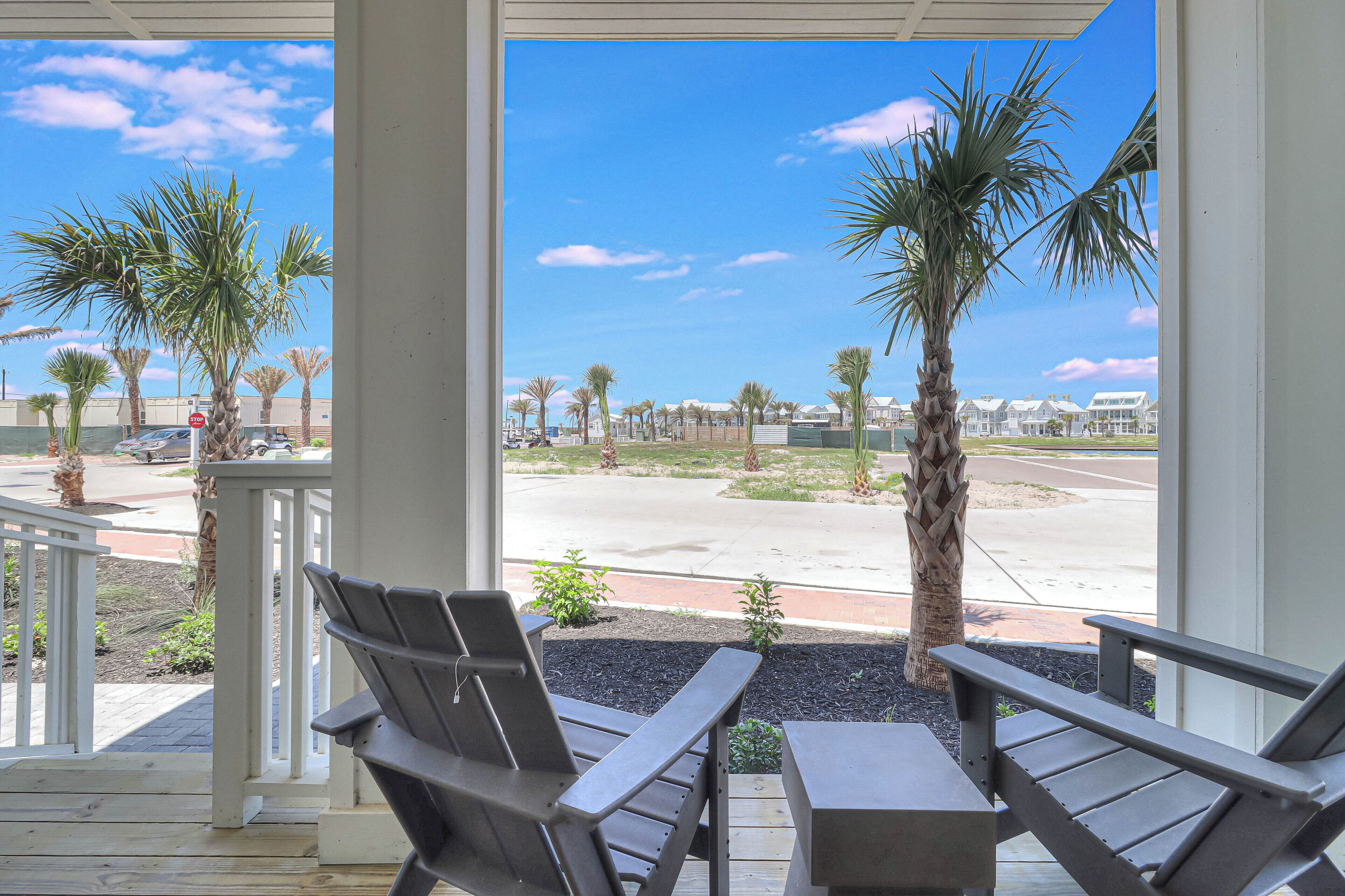 A covered outdoor seating area with two Adirondack chairs and a table, overlooking a parking lot and a row of palm trees.