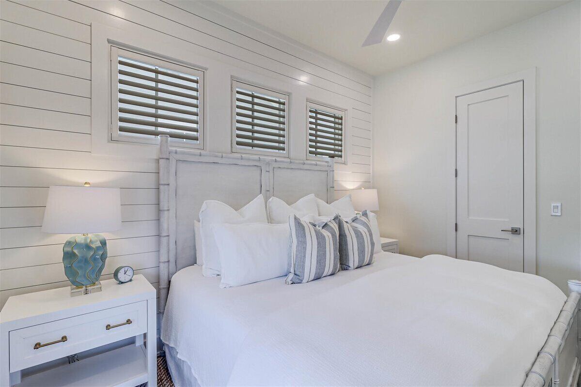 A bedroom with a large bed, white headboard, and three windows with blinds. There is a nightstand with a lamp and a clock.