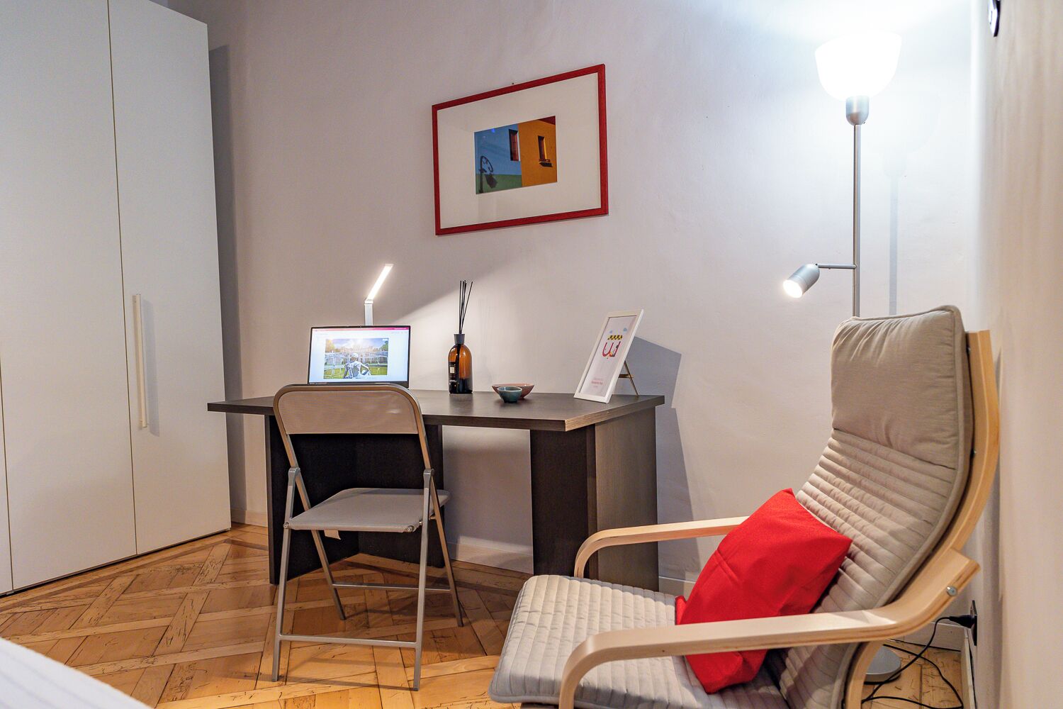 Een kleine kamer met een bureau, twee stoelen, een laptop, een ingelijste foto en een lamp.