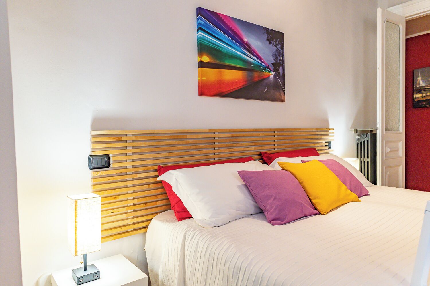A neatly made bed with a wooden headboard, a bedside table with a lamp, and a colorful painting above the bed.