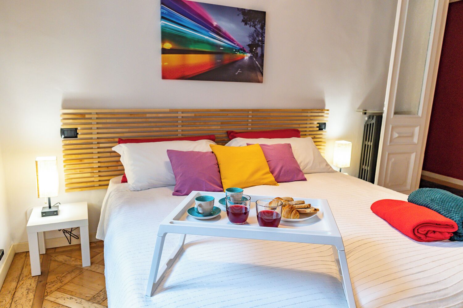 A hotel room with a bed, bedside tables, a tray with breakfast items, and a painting on the wall.