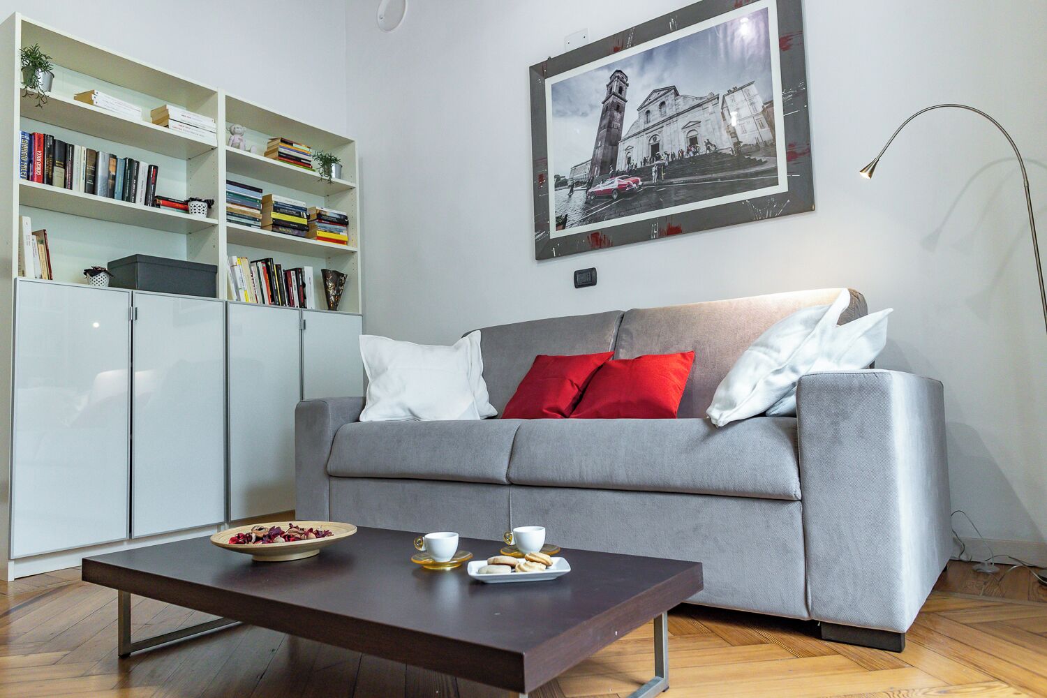 En moderne stue med en grå sofa, en bogreol, et billede i ramme på væggen og et sofabord med kopper og en tallerken.