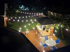 Un patio con mesa y sillas, un rincón cubierto para sentarse y luces colgantes.