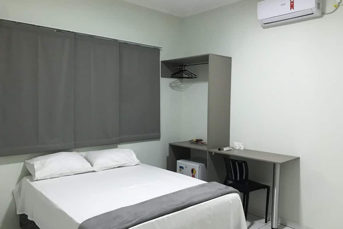 A bedroom with a bed, a desk, a chair, a small fridge, and an air conditioning unit.