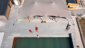 An aerial view of a swimming pool area with a wooden deck, lounge chairs, and a few people.