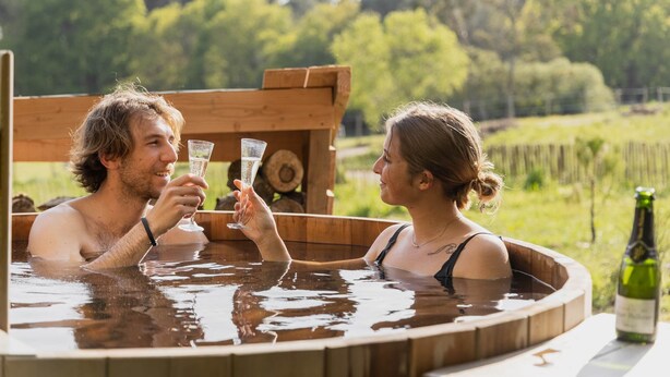 Two people in a wooden hot tub with a bottle of champagne on a table.